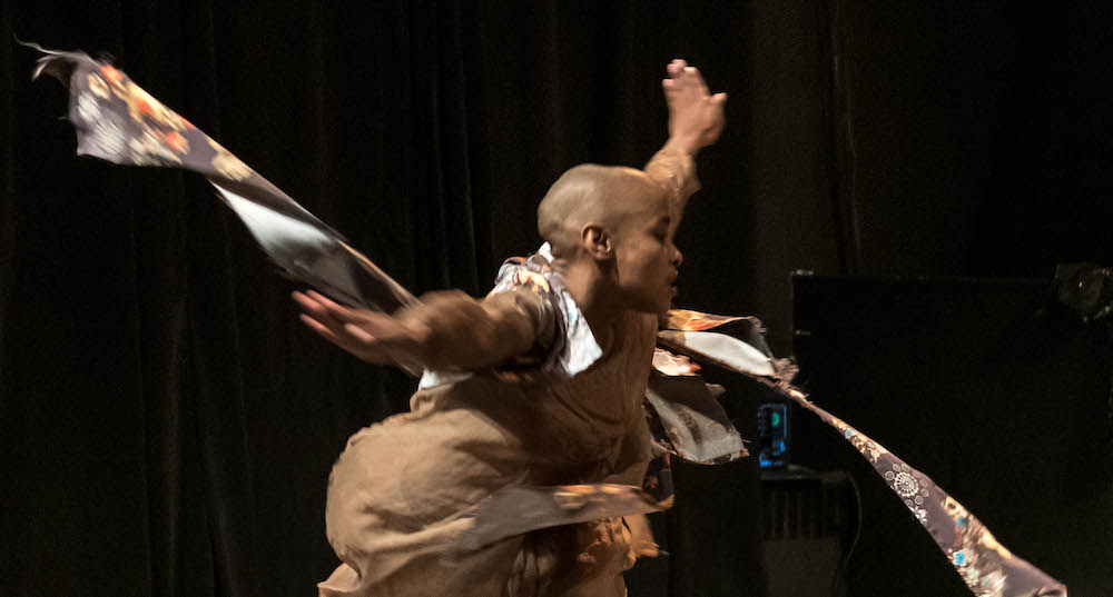 Jasmine hearn,  a light skinned bald Black woman in mid swirl . An upper body shot showing her costuming flying in the air as she turns. 